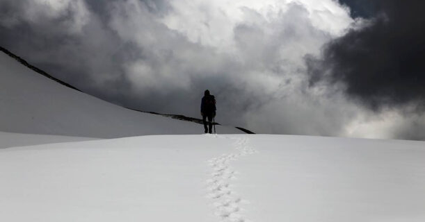 A hiker trekking through snow on a mountain under a cloudy sky.