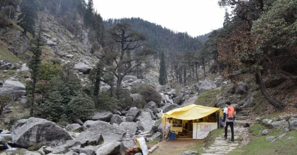 A hiker with an orange backpack walks along a rocky path toward a yellow tent in a mountainous area.