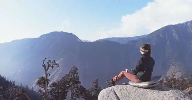 A person sits on a large rock, gazing at a panoramic view of distant mountain ranges under a clear sky.