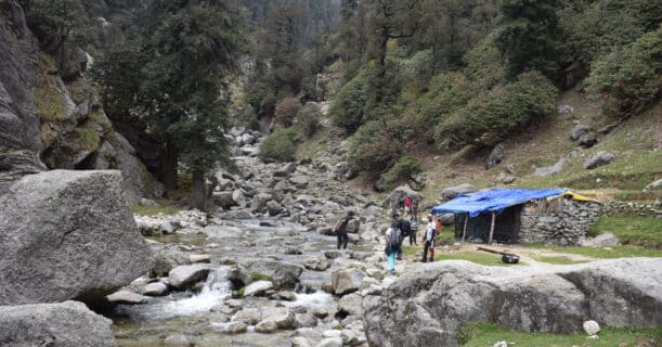 Hikers navigate a rocky stream in a mountainous forest with a blue tarp shelter in the background.