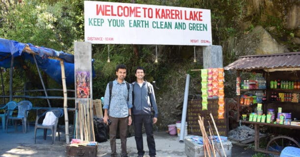 Two hikers standing at the entrance of Kareri Lake, surrounded by trekking gear and a sign promoting environmental cleanliness.