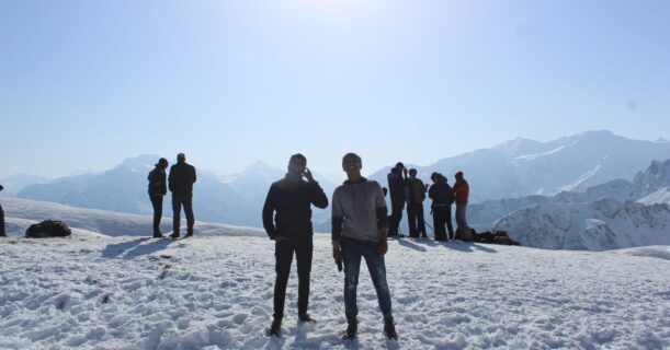 A cluster of people standing on a snowy mountain peak with the sun shining in the background.