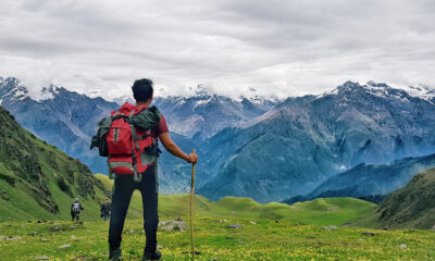 A man with a backpack standing on a grassy hill with mountains in the background.