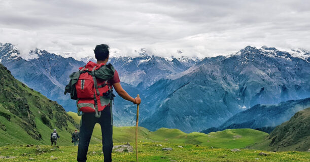 A man with a backpack standing on a grassy hill with mountains in the background.