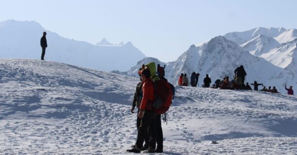 Individuals celebrate on a snowy mountain peak, with additional people in the background, capturing the beauty of the landscape.