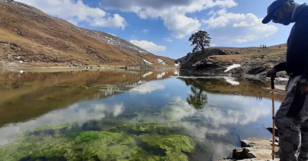 Trekker standing beside Brahmatal Lake reflecting mountains and clouds in Uttarakhand