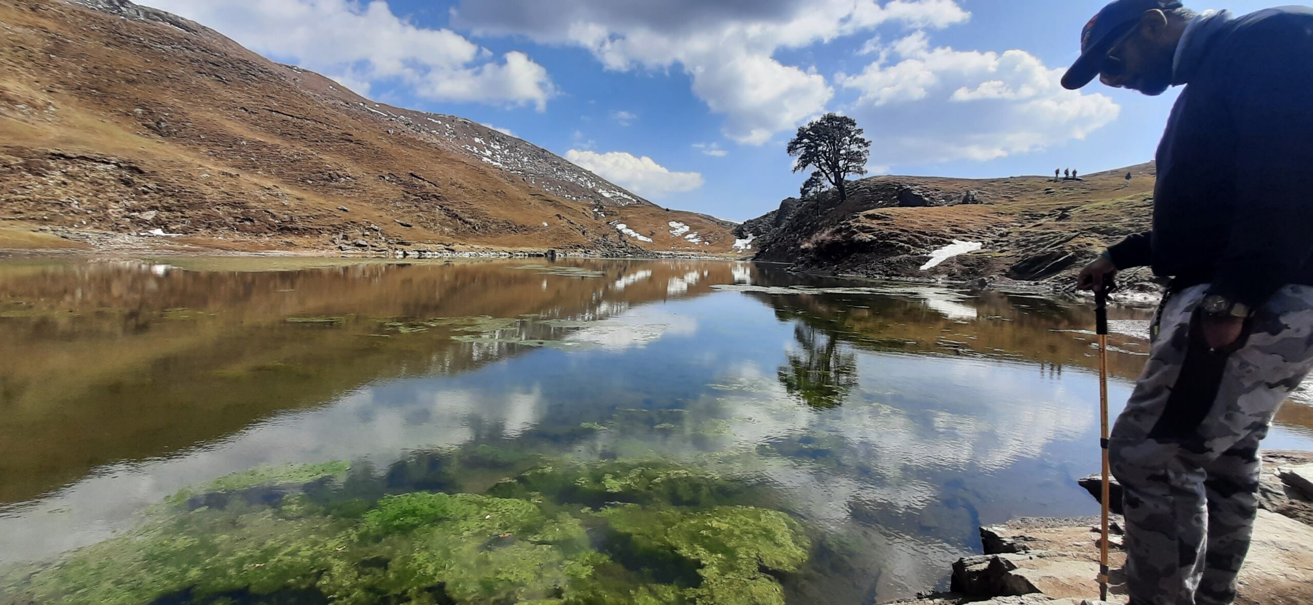 Trekker standing beside Brahmatal Lake reflecting mountains and clouds in Uttarakhand