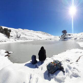 Trekker sitting by the frozen Brahmatal Lake under bright winter sun in Uttarakhand