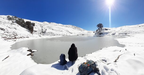 Trekker sitting by the frozen Brahmatal Lake under bright winter sun in Uttarakhand