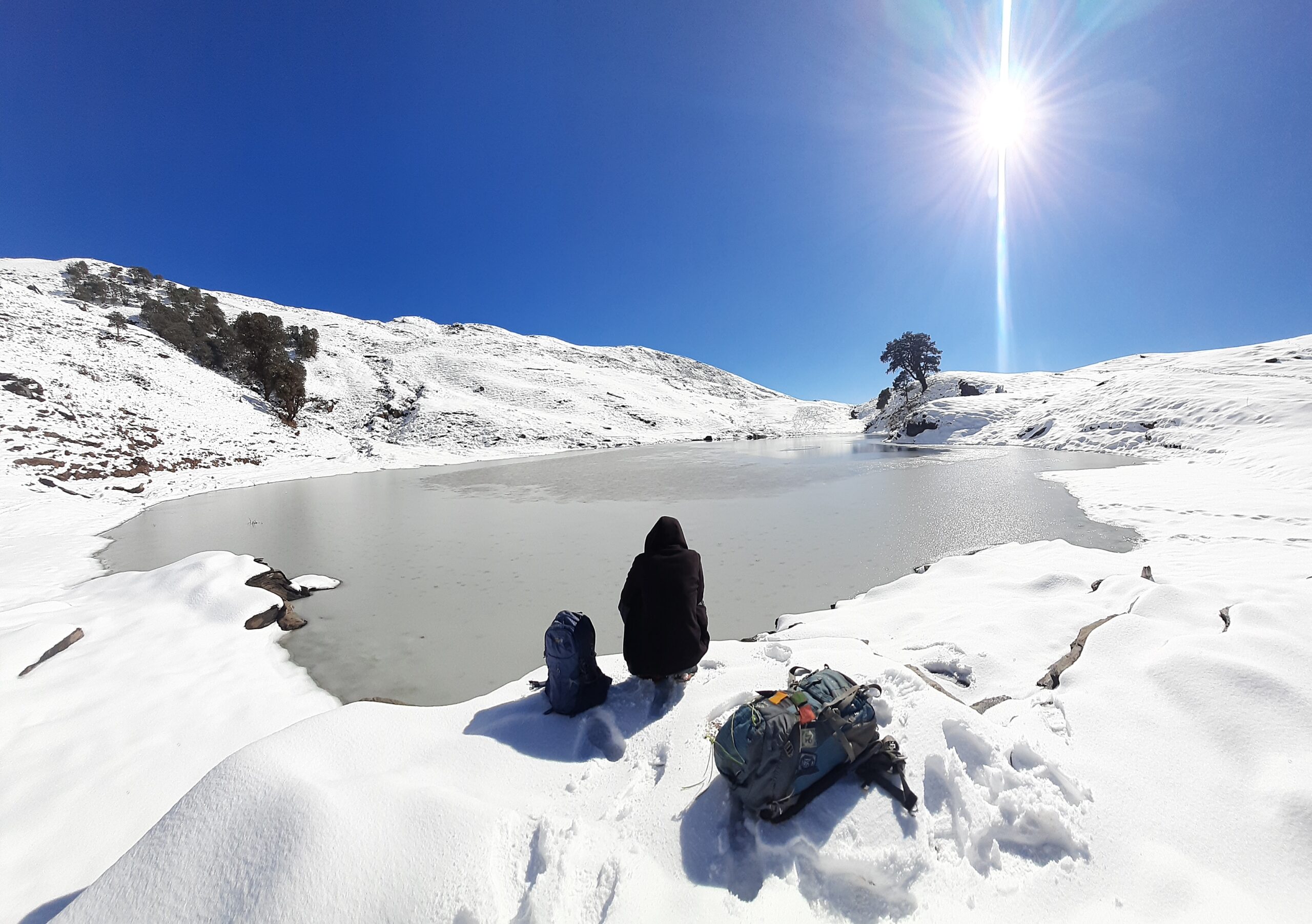 Trekker sitting by the frozen Brahmatal Lake under bright winter sun in Uttarakhand