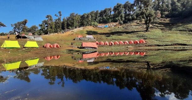 Colorful Brahmatal trek campsite reflected in crystal clear water under blue sky
