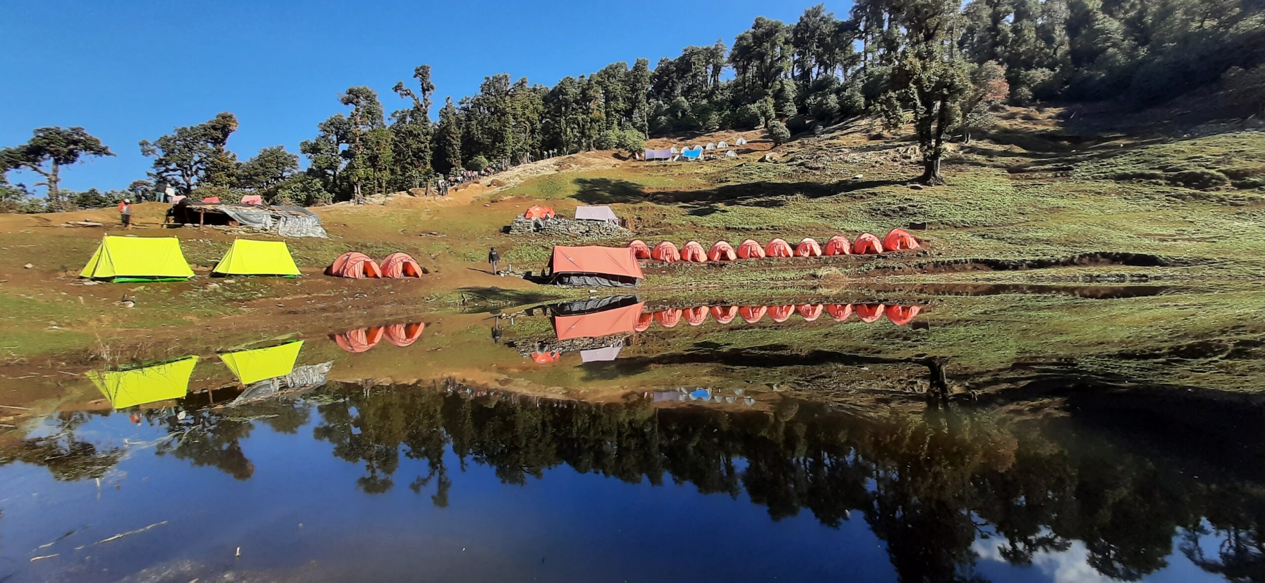Colorful Brahmatal trek campsite reflected in crystal clear water under blue sky