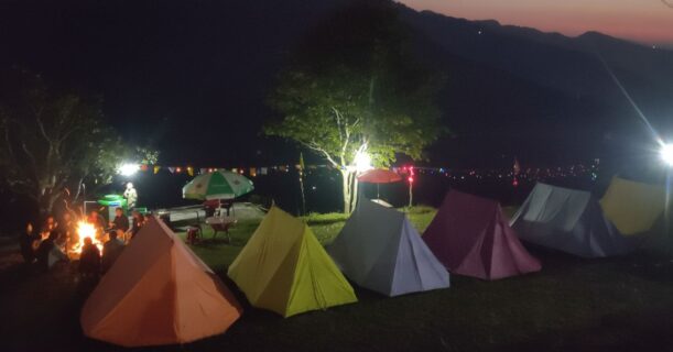 Colorful tents and trekkers enjoying a warm bonfire at Nag Tibba campsite under a clear night sky.