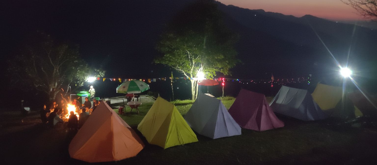 Colorful tents and trekkers enjoying a warm bonfire at Nag Tibba campsite under a clear night sky.