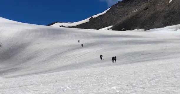 Trekkers climbing a snow-covered slope toward Pin Parvati Pass under a deep blue sky.
