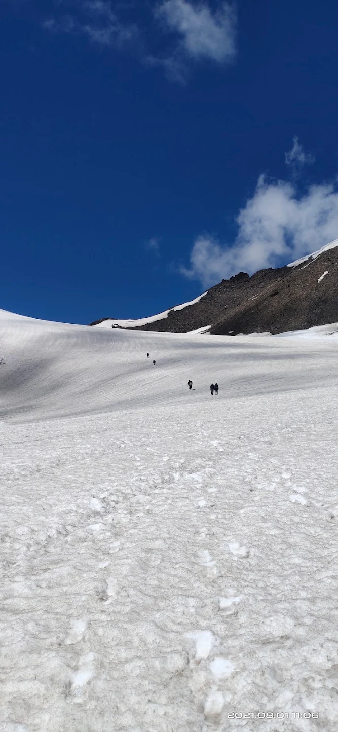 Trekkers climbing a snow-covered slope toward Pin Parvati Pass under a deep blue sky.