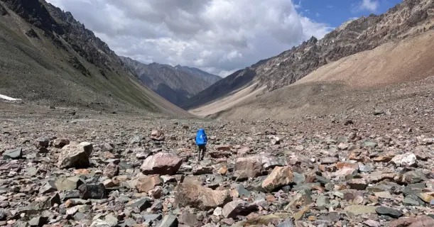 A trekker with a blue backpack walks across a vast, rocky valley in Spiti after descending Pin Bhabha Pass.