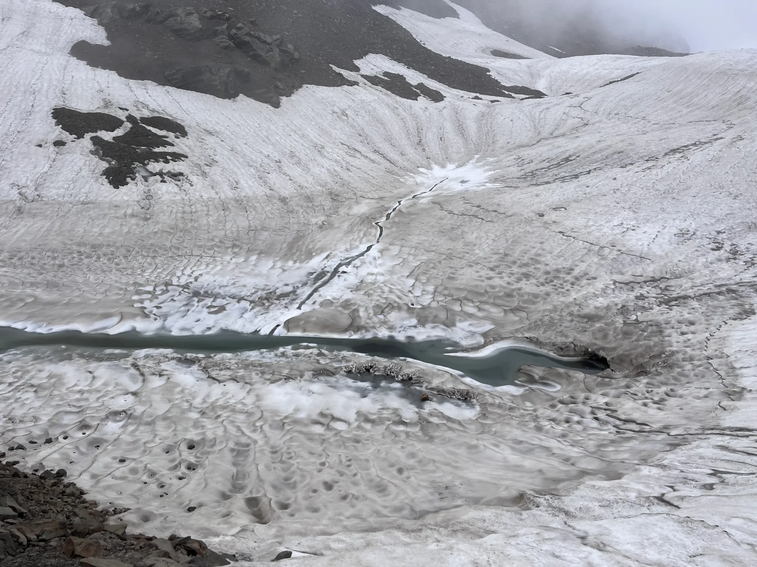 A high-altitude glacier field with a flowing meltwater stream and crevasses on the Pin Bhabha Pass trail.