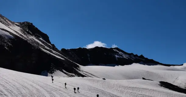 Trekkers walking across the snow-covered glacier near Pin Parvati Pass under a bright sun and clear blue sky.