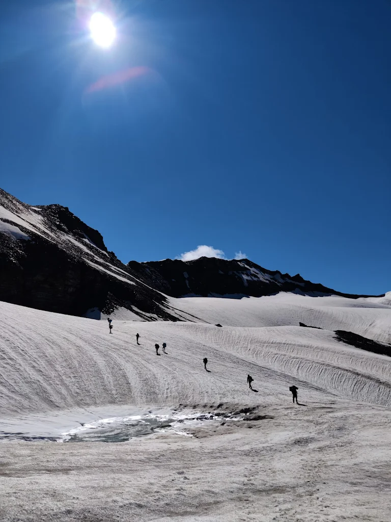 Trekkers walking across the snow-covered glacier near Pin Parvati Pass under a bright sun and clear blue sky.