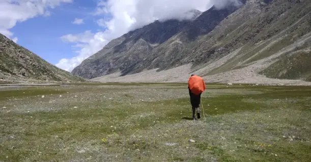 Trekker with an orange backpack cover walking through a wide alpine meadow surrounded by mountains on the Pin Parvati trek.
