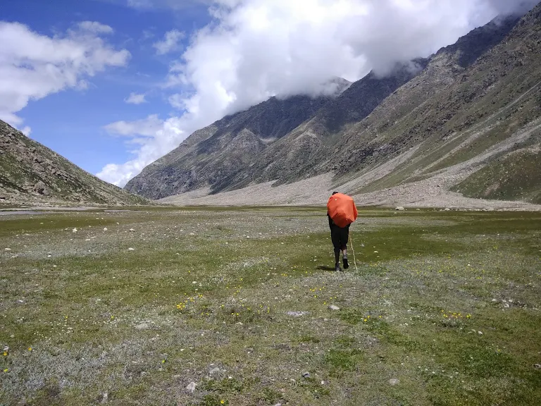 Trekker with an orange backpack cover walking through a wide alpine meadow surrounded by mountains on the Pin Parvati trek.