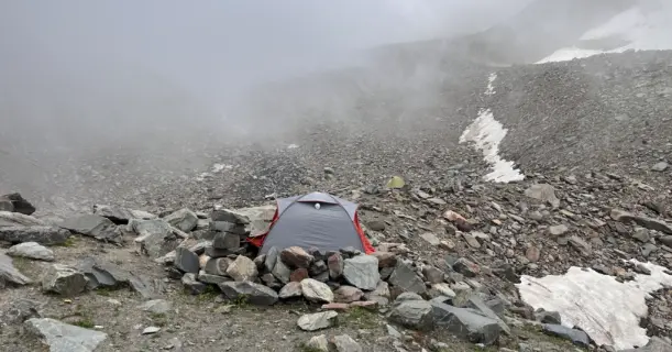 A trekking tent pitched among rocks on the high-altitude, barren terrain of Pin Bhabha Pass with mist and snow patches in the background.