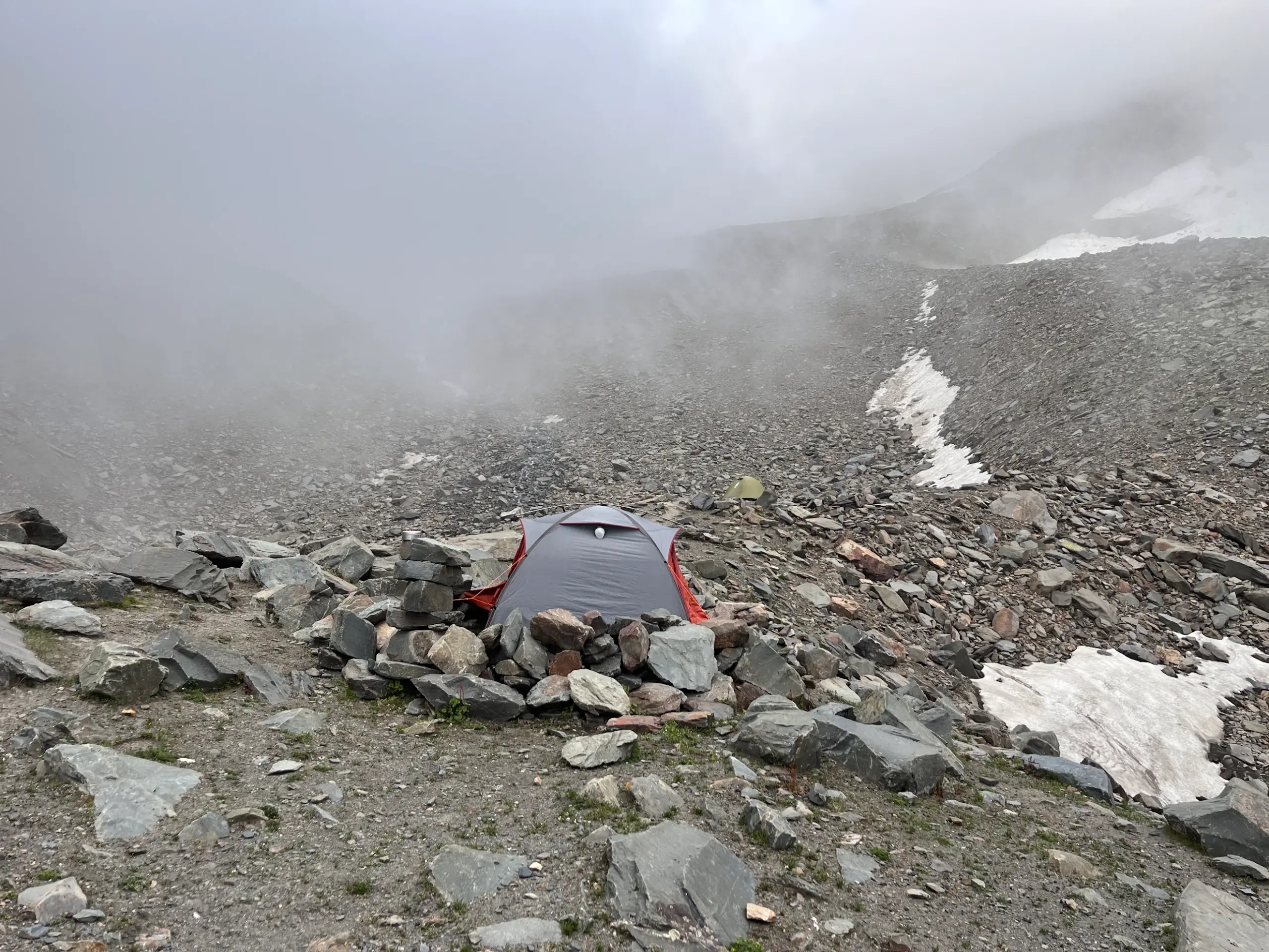 A trekking tent pitched among rocks on the high-altitude, barren terrain of Pin Bhabha Pass with mist and snow patches in the background.