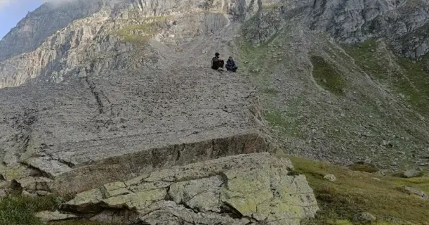 Trekkers sitting on a large natural rock slab with mist-covered mountains behind, near Pandu Pul on the Pin Parvati trek.
