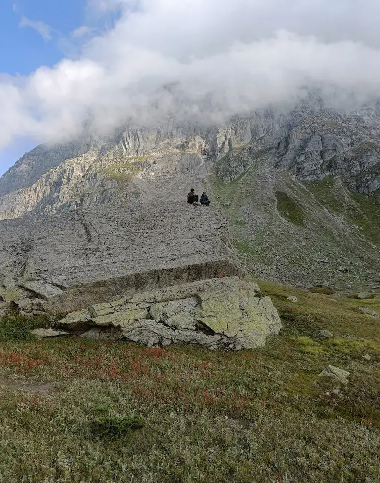 Trekkers sitting on a large natural rock slab with mist-covered mountains behind, near Pandu Pul on the Pin Parvati trek.