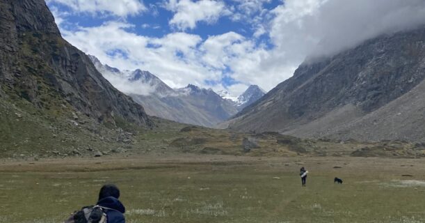 Trekker walking through a wide alpine meadow on the Pin Parvati Pass trek, with mountains in the distance under a partly cloudy sky.