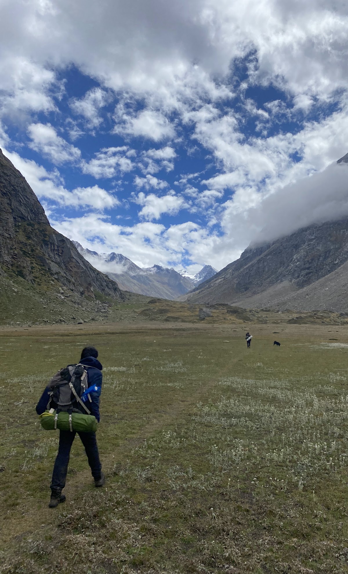 Trekker walking through a wide alpine meadow on the Pin Parvati Pass trek, with mountains in the distance under a partly cloudy sky.