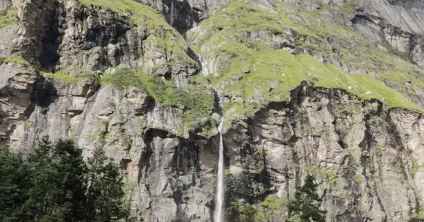 Tall waterfall cascading down a lush green cliff with a rainbow at the base on the Pin Parvati Pass trek.