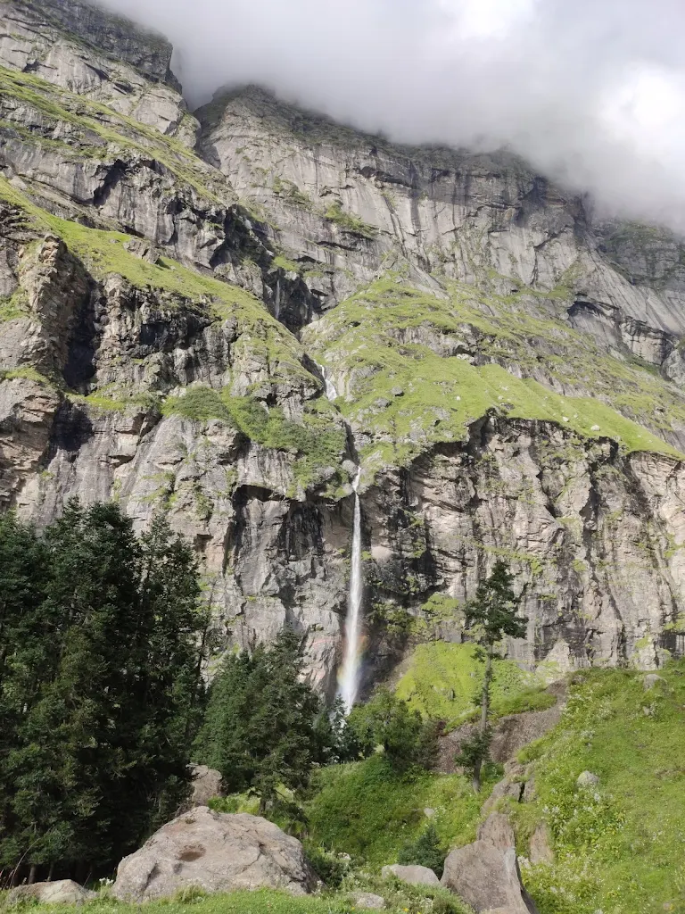 Tall waterfall cascading down a lush green cliff with a rainbow at the base on the Pin Parvati Pass trek.