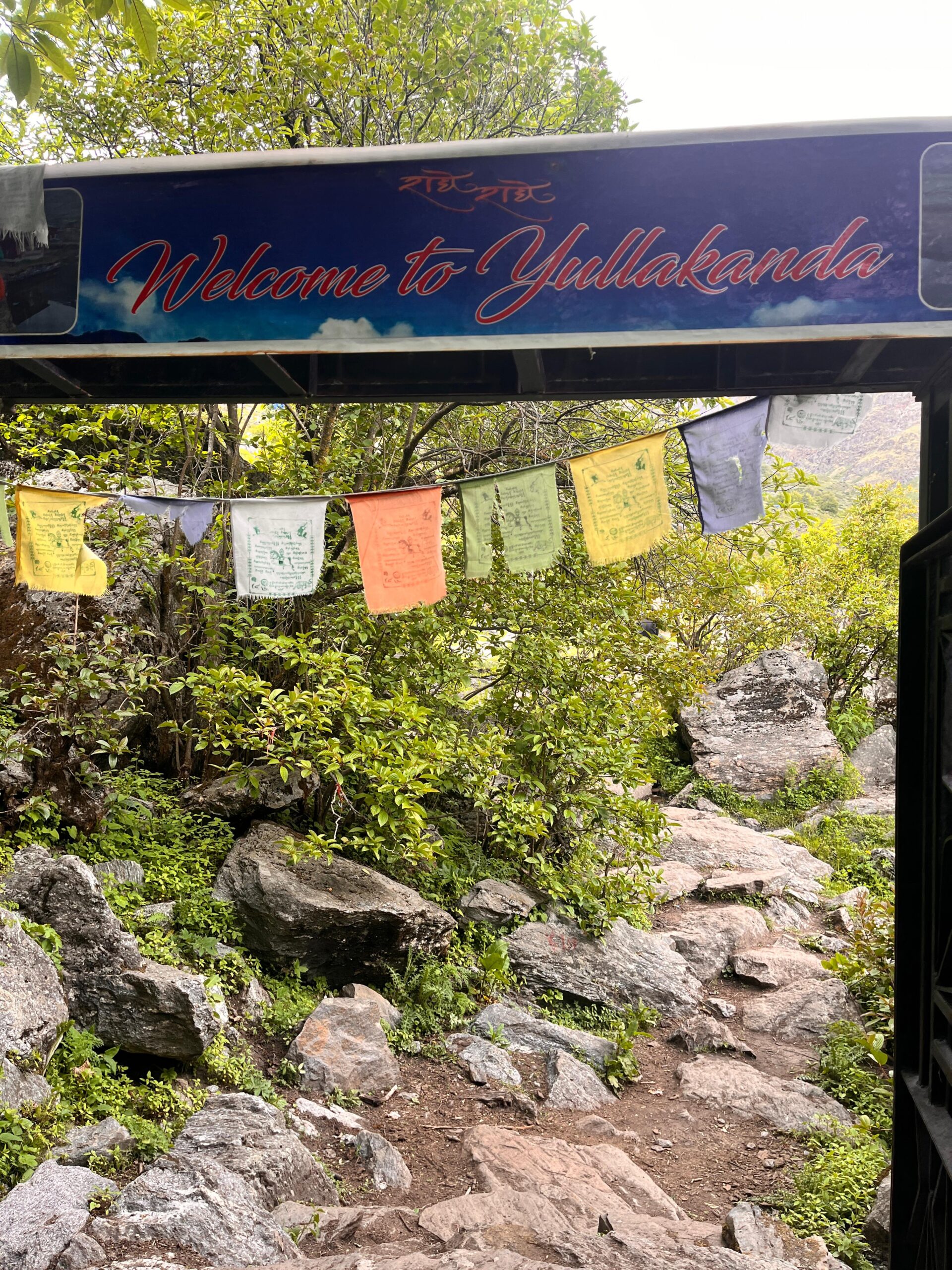 Entrance signboard reading “Welcome to Yulla Kanda” with colorful prayer flags and rocky trekking trail in Himachal Pradesh
