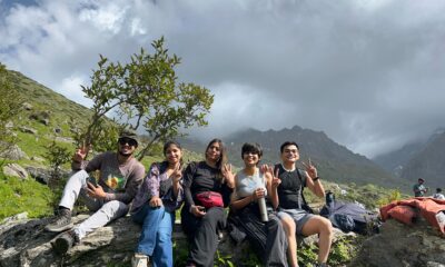 Group of young trekkers resting on rocks with lush green meadows and misty Himalayan peaks at Yulla Kanda trek