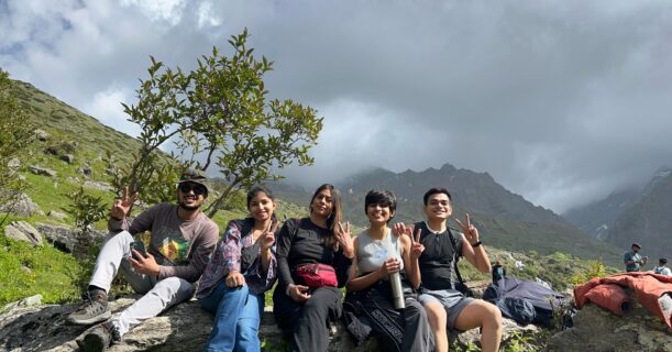 Group of young trekkers resting on rocks with lush green meadows and misty Himalayan peaks at Yulla Kanda trek