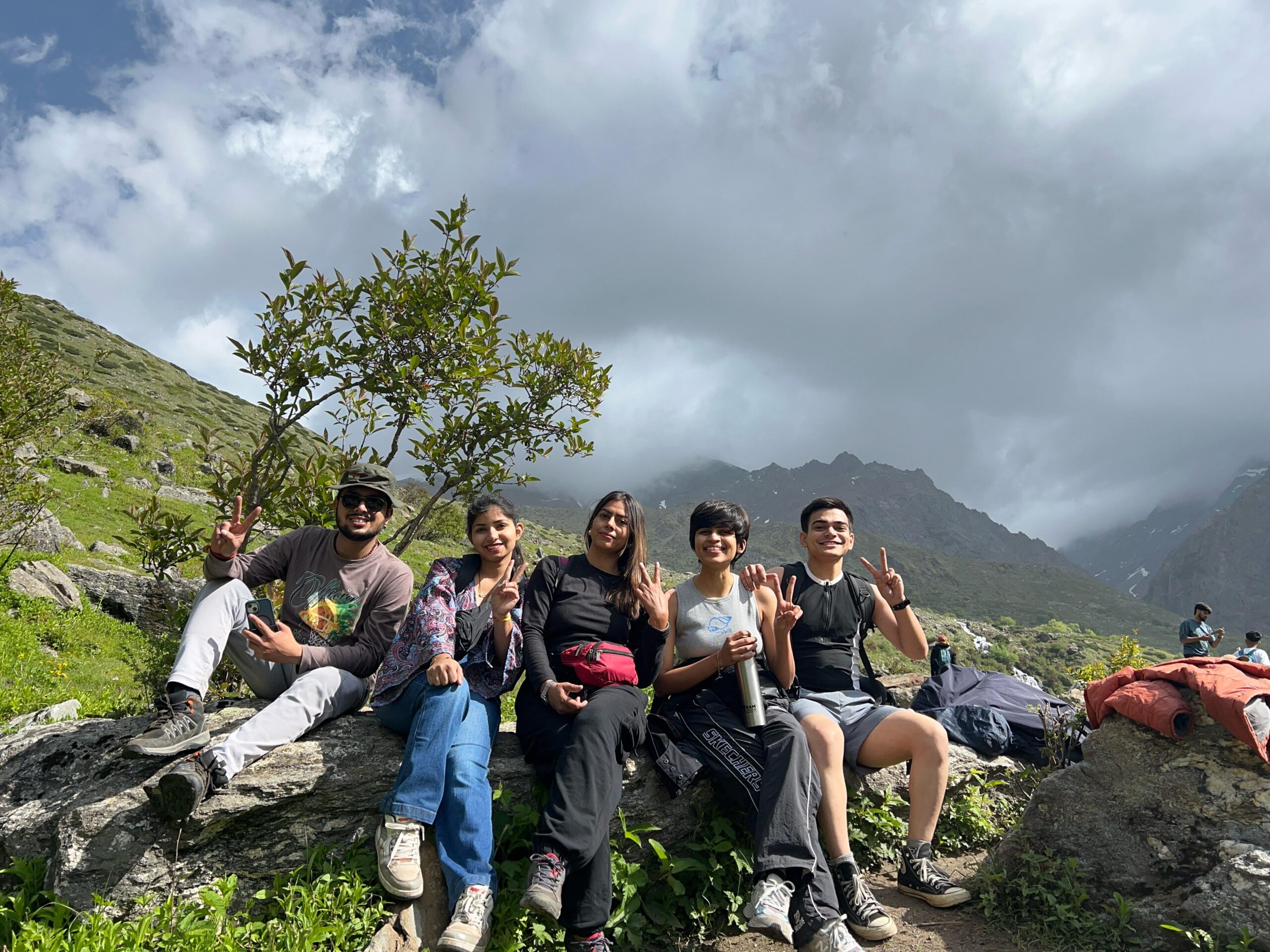 Group of young trekkers resting on rocks with lush green meadows and misty Himalayan peaks at Yulla Kanda trek